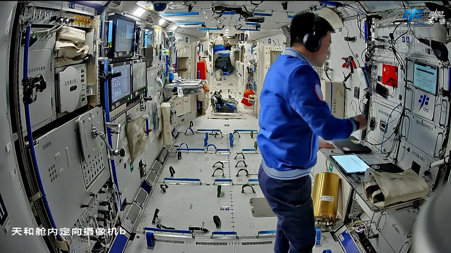 A Chinese astronaut wearing headphones moves his right hand above a tablet inside the Tianhe core module of the China Space Station. The original video watermark and camera label remain visible.