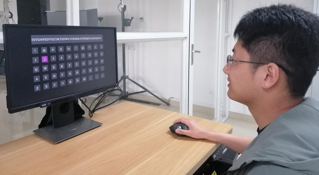 A participant uses gaze to point at an on-screen keyboard and a trackball button to confirm selections, with an eye tracker mounted below the monitor.